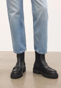 Svarta klumpiga läderskor med ankeln kända för att bäras med ljusblå raka jeans på en polerad betonggolv.