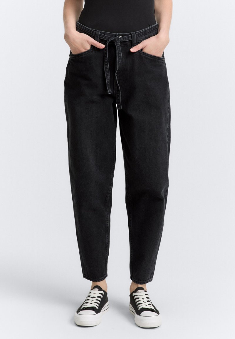 Pantalones de mezclilla negros con cintura alta, corte relajado, dos bolsillos frontales y un cinturón de tela. Combinados con zapatillas de lona negras.
