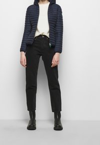 Henkilö, joka pitää yllään tummansinistä tikattua huppujakkua, valkoista neuletta, mustia korkeavyötäröisiä farkkuja ja mustia nilkkureita, seisoo yksinkertaista taustaa vasten.