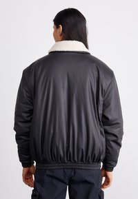 Chaqueta bomber negra de material sintético con un cuello de forro polar crema, cinturilla y puños elásticos, de textura suave y diseño minimalista.