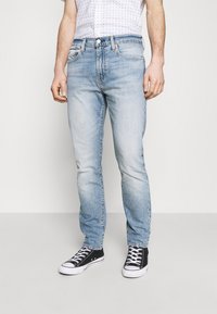 Vaqueros de mezclilla azul claro con corte recto, que presentan un diseño clásico de cinco bolsillos, costuras en contraste y cremallera frontal. Combinados con zapatillas negras.