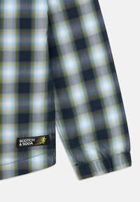 Prugasta košulja u plavoj, zelenoj i bijeloj boji s mekim tekstilnim uzorkom, kopčanje na tipke i crna oznaka s natpisom "SCOTCH & SODA" na rubu.