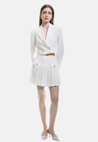 Witte cropped blazer met dubbele knopen, gecombineerd met een geplooide witte minirok met twee voorzakken en metalen accenten, gedragen met hakken.