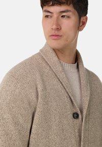 Ung man som vänder huvudet åt sidan, iförd en beige strukturerad kofta över en ljusstickad tröja framför en vit bakgrund.