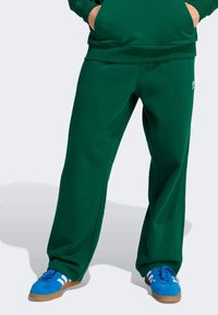 Zielone dresy z miękkiego materiału, w luźnym kroju, z bocznymi kieszeniami i małym logo na lewym udzie, połączone z niebieskimi butami.