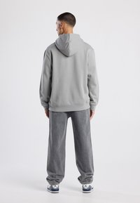 Uomo che indossa un felpa con cappuccio grigia, jeans larghi grigi e sneakers grigie e bianche, in piedi con la schiena rivolta verso uno sfondo neutro.