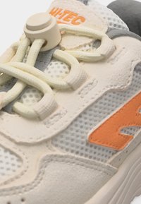 Közelkép egy bézs és szürke sportcipőről narancssárga logóval, hálós anyaggal, műbőr rávarrásokkal és egyedi csúszkás fűzőrendszerrel.