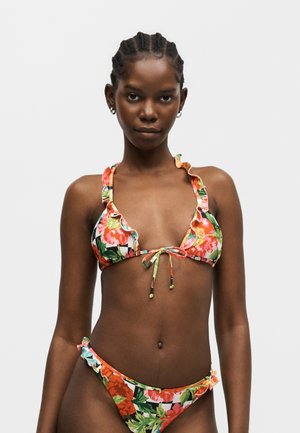 Jauna moteris su austomis plaukais, vilkinti spalvotus gėlių rašto bikinį su raukiniais, pozuojanti prieš paprastą šviesų foną.