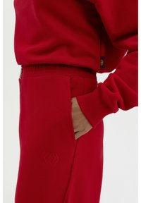 Roter Sweatshirt mit passenden Jogginghosen, ausgestattet mit einem gerippten Bündchen, Seitentasche und gesticktem Logo-Detail auf den Jogginghosen. Weicher Stoff.