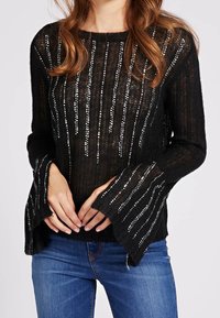Pull en maille noir avec des rayures en sequins argentés, des manches cloche et un col rond. Associé à un jean bleu, il présente un design loose et texturé.