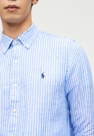 Άνδρας που φοράει πουκάμισο με κατακόρυφες ριγέ σε ανοιχτό γαλάζιο και άσπρο, με λογότυπο παίκτη πόλο σε ναυτικό μπλε στο στήθος.