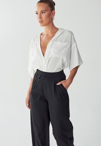 Witte kortekleurige shirt met een diepe V-hals en twee borstzakken, gecombineerd met hoge zwarte broeken met zijzakken en plooien.