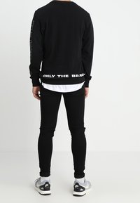 Sudadera negra con texto blanco en el dobladillo y mangas largas, combinada con vaqueros ajustados negros y zapatillas grises. Textura de tela lisa.