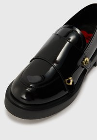 Mocassino in pelle patent nera con suola spessa, dotato di dettagli a forma di cuore in oro e una finitura liscia e lucida. Design a punta rotonda.