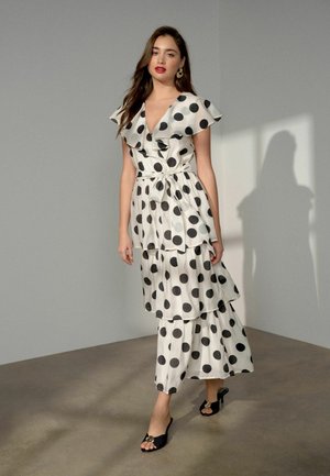 Frau steht drinnen und trägt ein weißes Maxikleid mit großen schwarzen Punkten, Rüschensäumen, einem Taillenband und schwarzen offenen Absatzschuhen.