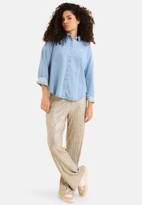 Camicia di jeans azzurro chiaro con maniche risvoltate, chiusura a bottoni e dettagli ricamati dorati, abbinata a pantaloni argentati lucidi e plissettati.