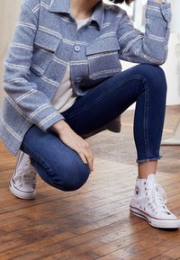 En person som hukar sig inomhus och bär en blå rutig jacka, mörka skinny jeans och vita högskaftade Converse-skor på trägolvet.