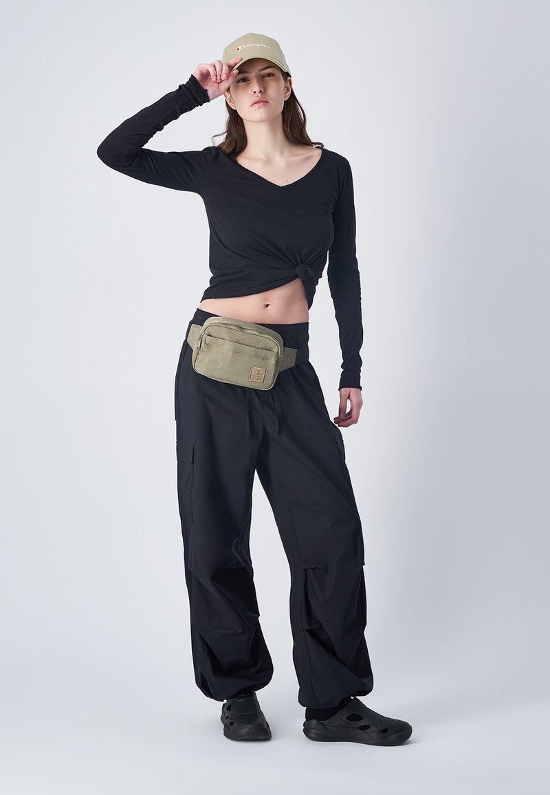 Maglietta nera a maniche lunghe annodata in vita, pantaloni cargo neri larghi, marsupio verde oliva e calzature nere, con un cappello beige.