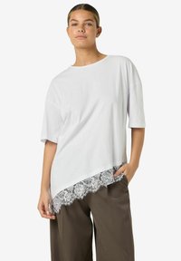 Hvit oversized t-skjorte med en buet kant og en bølget blonderand. Stoffet ser glatt ut, kombinert med løse olivengrønne bukser.