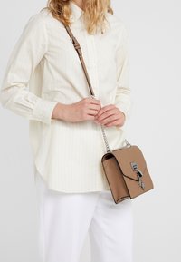 Brązowa skórzana torba na ramię z srebrnymi akcentami, prostokątny kształt i regulowany pasek. Posiada zapięcie na klapę oraz subtelne teksturowane wykończenie.