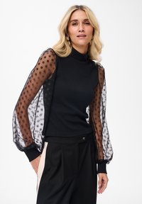 Czarny top z dopasowanym żeberkowym gorsetem i przezroczystymi, wzorzystymi rękawami z czarnymi kropkami. Zestawiony z czarnymi spodniami, ukazując elegancką sylwetkę.