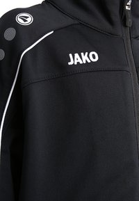 Zwarte ritsshirt gemaakt van zachte stof, met witte afwerking en stippen op de schouders. De merknaam "JAKO" is prominent in het wit afgebeeld.