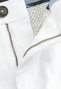 Pantaloni corti bianchi in tessuto con chiusura a zip e accessori metallici marroni, dotati di una fodera interna a motivo di piccole forme geometriche.