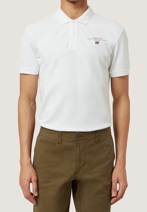 Mężczyzna ubrany w biały polo z krótkim rękawem z logo Napapijri i małą norweską flagą, w zestawieniu z oliwkowozielonymi spodniami.