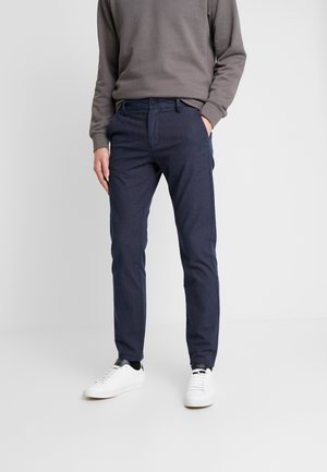Person trägt marineblaue Slim-Fit-Hose, graues Langarmshirt und weiße Sneakers, steht mit einer Hand in der Tasche vor weißem Hintergrund.