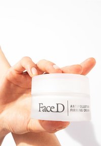 Barattolo bianco opaco con coperchio trasparente, etichettato "FaceD ANTIPOLLUTION FIRMING CREAM", tenuto in una mano. Texture liscia, forma rotonda.