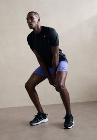 Schwarzes sportliches T-Shirt, lila Shorts und schwarze Turnschuhe. Die Person hält eine Kettlebell und hat eine konzentrierte Körperhaltung in einer neutralen Umgebung.