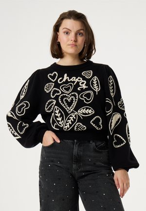Schwarzer Pullover mit weißen gestickten Herzen und Blättern, runder Ausschnitt und langem Puffärmel. Getragen mit hochgeschnittenen dunklen Jeans.