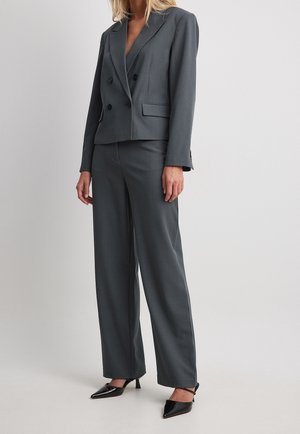 Femme portant un blazer gris foncé croisé avec un pantalon large assorti et des talons noirs à bout pointu.