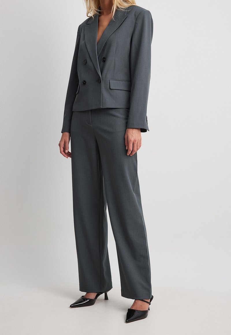 Femme portant un blazer gris foncé croisé avec un pantalon large assorti et des talons noirs à bout pointu.