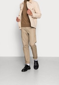 Ljusbeige jacka med bruna detaljer, brun polotröja, beige byxor och svarta skor. Vita strumpor är synliga och har en slät textur.