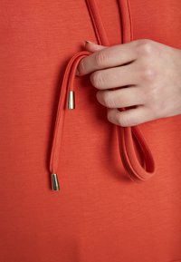 Oranges Sweatshirtstoff mit glatter Textur, ausgestattet mit einem Kordelzug mit goldenen Metallenden, der in einer Hand gehalten wird.