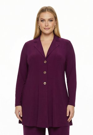 Frau mit langen blonden Haaren trägt einen langen violetten Blazer mit Knöpfen und Revers sowie passende Hose vor weißem Hintergrund.