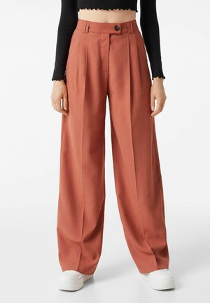 Bredbenede rust-orange bukser med front plisseringer, knap- og lynlåslukning, båret med hvide sneakers og en sort cropped langærmet top.