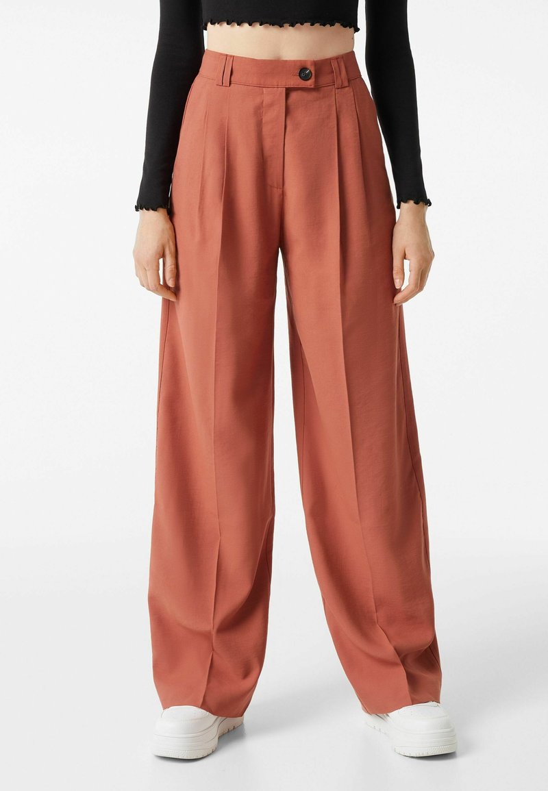 Pantalon large orange rouille avec plis sur le devant, fermeture par bouton et zip, porté avec des baskets blanches et un top court noir à manches longues.