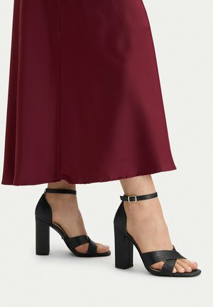 Piedi che indossano sandali neri con tacco alto e cinturini alla caviglia abbinati a una gonna midi bordeaux su uno sfondo chiaro.