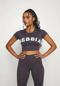 Αθλητική γυναίκα με πλεξούδες στέκεται με τα χέρια στη μέση, φοράει ένα σκούρο κοντό τοπ με κείμενο "NEBBIA" και ταιριαστά ψηλόμεσα κολάν.