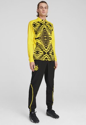Langærmet gul skjorte med sort zebra mønster, parret med sorte bukser med gule detaljer og et logo på siden.