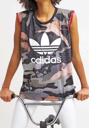 Osoba nosząca bezrękawnik Adidas z motywem żurawi i fal, trzymająca kierownicę roweru, ubrana w białe spodnie.