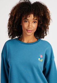 Modrá mikina s okrúhlym výstrihom a voľným strihom, ozdobená farebným logom na ľavej hrudi a hladkou, jemnou textúrou.