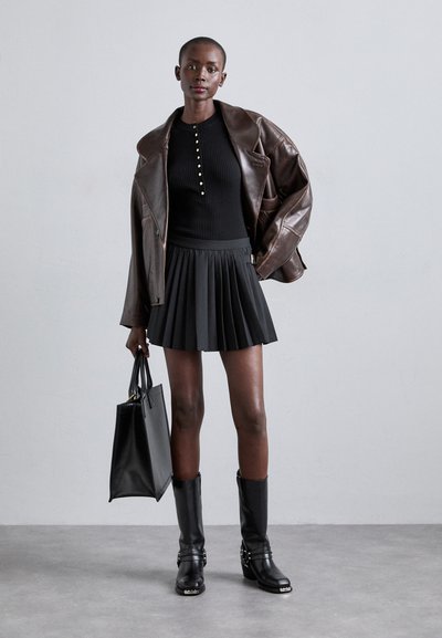 Schwarzes geripptes Oberteil mit goldenen Knöpfen, braune Lederjacke in Übergröße, schwarzer Falten-Minirock, schwarze kniehohe Stiefel und schwarze Handtasche.