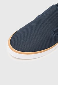 Mornarsko plava platnena cipele slip-on s teksturiranim gornjim dijelom, zaobljenim vrhom i gumenim potplatom boje gume s bijelim rubom. Sadrži minimalno šivanje.