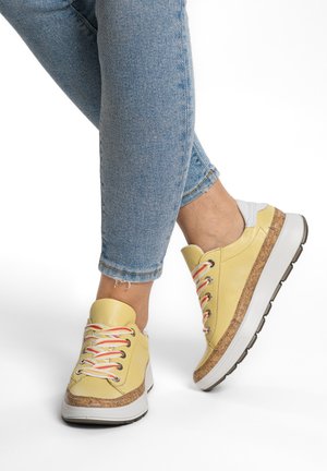 Person wearing light blue jeans and yellow sneakers with cork-textured trim and white soles, one foot slightly raised.