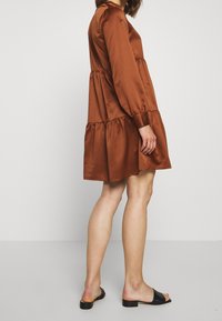 Brun satäng klänning med lång ärm, med lager på lager volangnederkant, samlad midja och rund halsringning. Matchad med svarta slidesandaler.