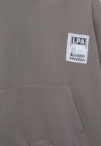 Grå hoodie med en texturerad velour, med en framficka och en vit etikett med svart text, placerad på vänster sida.