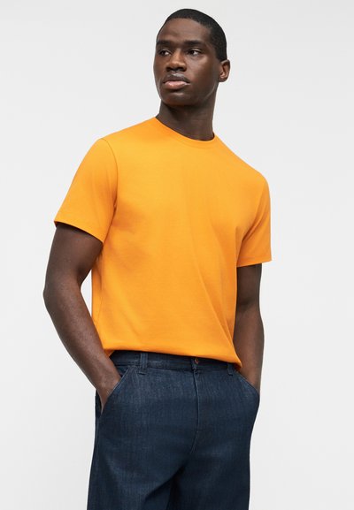 Άνδρας που φοράει φωτεινή πορτοκαλί κοντομάνικη μπλούζα και σκούρο μπλε τζιν, στέκεται με τα χέρια στις τσέπες, κοιτάζοντας προς τα πλάγια σε ένα απλό φόντο.